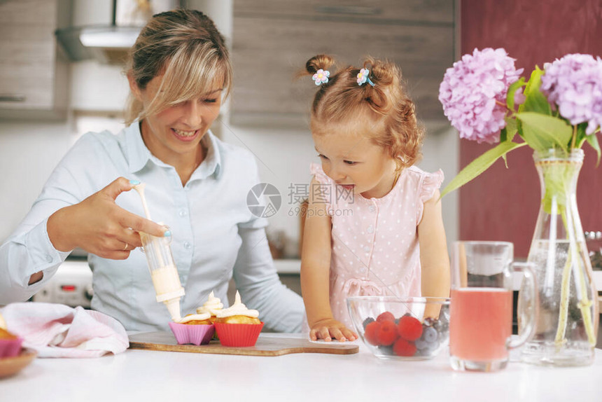 一个漂亮的小女孩和她的小女儿用奶油装饰新鲜出炉的纸杯蛋糕桌上有纸杯蛋糕浆果汁图片