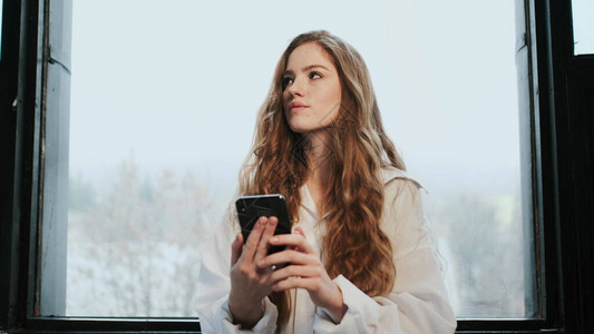 长头发的年轻女孩站在窗边手里拿着手机的女孩她考虑某图片