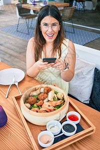 年轻漂亮的西班牙裔女人开心地笑着坐在桌子上用智能手机在餐厅图片