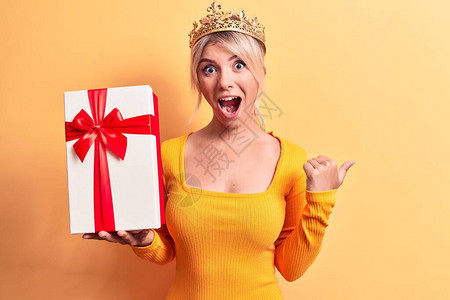 身戴金公主王冠的年轻金发美女拿着生日礼物举起大拇指向侧面图片
