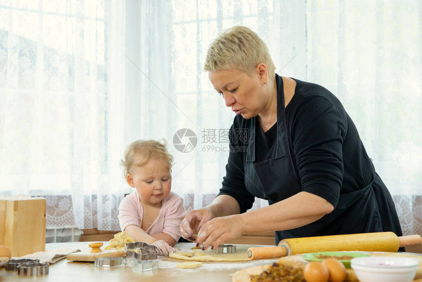 穿着黑色围裙的祖母教她可爱的小孙女如何制作姜饼小女孩和祖母在厨房的木桌上一起做饭幸图片