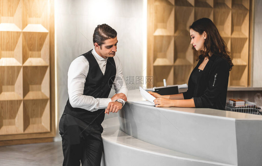 年轻英俊的商人与站在现代酒店大堂柜台接待处的女接待员交谈并图片