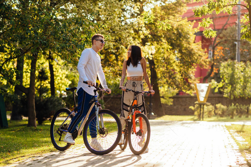 穿着时尚休闲服的快乐年轻人骑着自行车穿过美丽的秋季公园恩爱的年轻夫妇在日落的背景下在户外骑自行车度图片