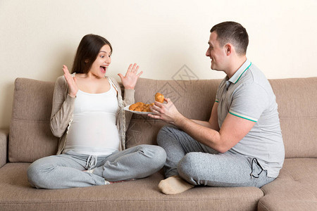 快乐的丈夫正在家里的沙发上用羊角面包喂他怀孕的妻子未来的父图片