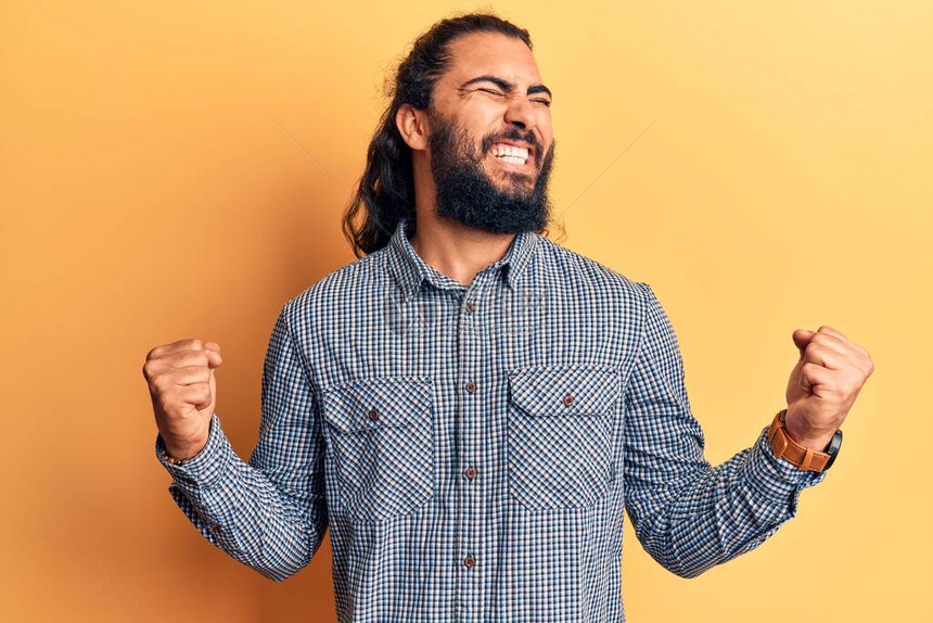 身着散装衣的阿拉伯青年男子非常高兴和奋地以举起手臂微笑和为成功而尖叫的方式做赢家姿图片