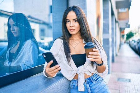 使用智能手机和饮酒严肃表达的年轻西班牙女孩在城市喝咖啡图片