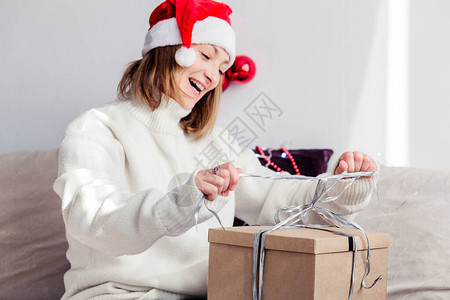 一个穿白色毛衣的年轻女子圣诞老人帽子在早上打开新年礼物横向图片