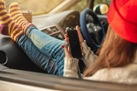 穿着温暖袜子的汽车中的女人在日落时手持机小装置和咖啡杯图片