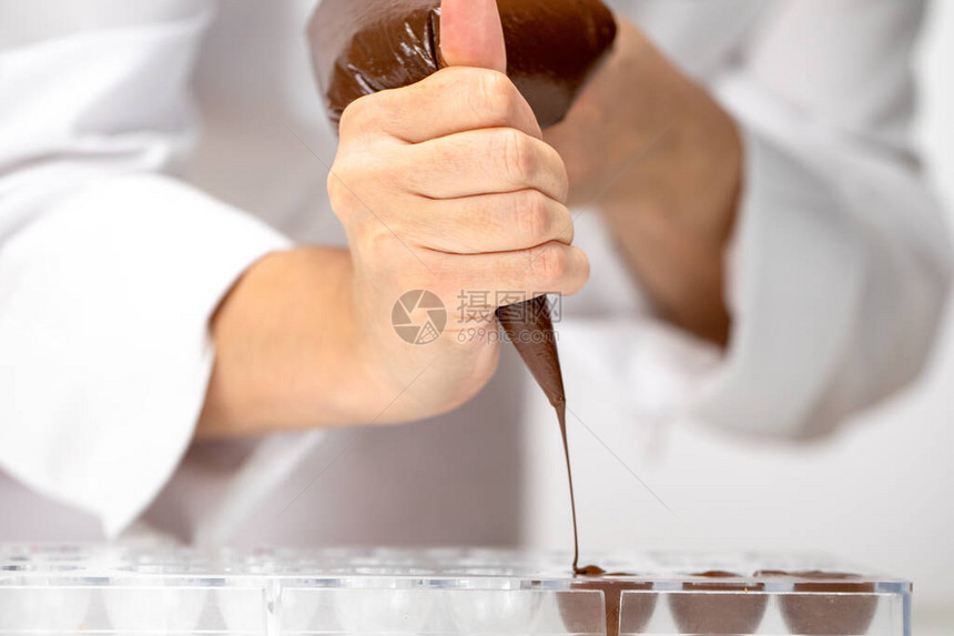 特写巧克力师将黑巧克力倒入糖果模具中厨师在白色围裙使用糕点袋填充塑料模具与热融化的巧克力制作自制巧克力糖图片