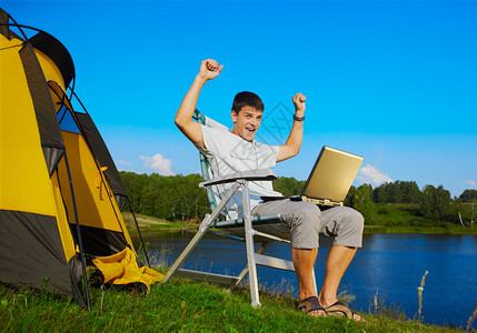 在露营帐篷户外附近坐在折叠椅上的手持笔记本电脑的辅助男子图片