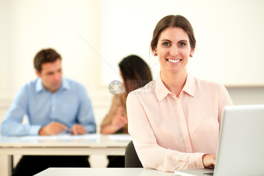 年轻的白人女在她的笔记本电脑上工作并坐在办公桌上的两个同事面前对图片