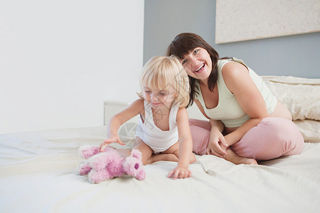 怀孕的母亲和蹒跚学步的小女儿坐在家里的床上图片