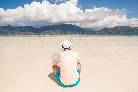 坐在沙滩上享受美丽的风景的年轻男人的后背戴着帽图片