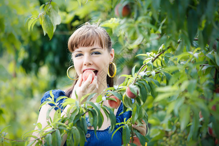 身着蓝裙子的美丽的黑发姑娘手里吃桃子站在绿色花园的笑图片