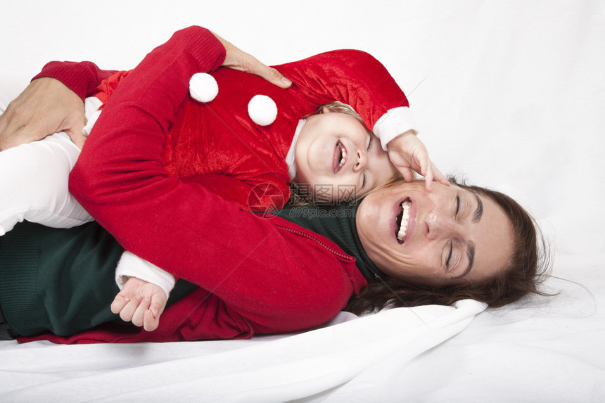 一岁高加索金发可爱婴儿圣诞老人的温柔而有趣的肖像与黑发女人母亲红色开衫绿色毛衣的圣诞伪装拥抱在一起笑着躺在白色图片