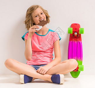 带着滑板吃巧克力的年轻女孩坐在工作室的地板上身着白图片