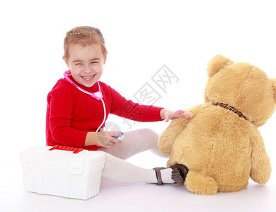 穿着红毛衣微笑的小女孩坐在地上和医院玩耍Teddy女孩用白色背景对待图片