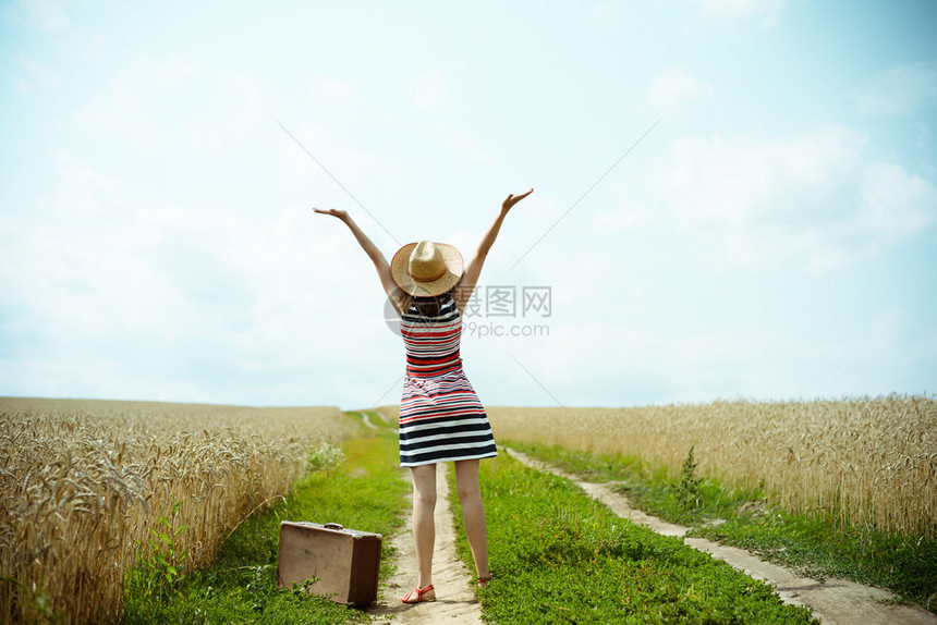 Backview带着旧手提箱在乡间小路上的幸福女人戴着帽子和条纹连衣裙的valize女孩举手到阳光明图片