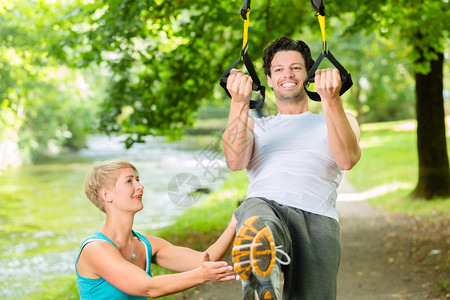 年轻的健身男子在城市公园的夏季树下用悬挂式教练吊索和私人图片