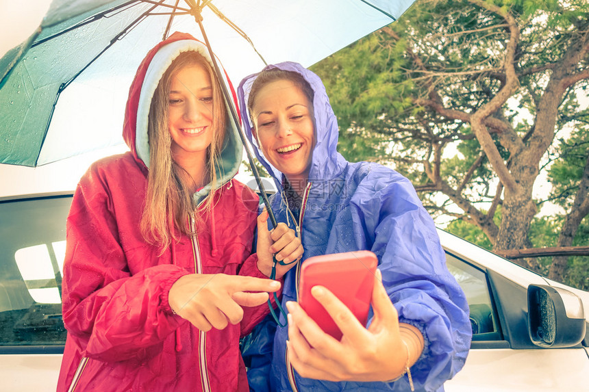 女最好的朋友在雨后阳光下享受智能手机与年轻女友一起玩的汽车旅行由于背光和阳光晕而具有柔和焦图片