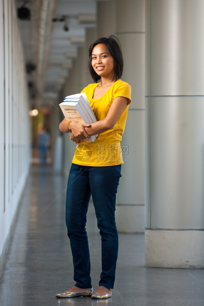全长肖像的微笑大学生站在走廊里拿着书看着相机的黄色衬衫图片