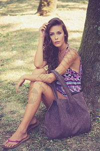 美丽的年轻女子穿着丁字裤和带皮包的夏装坐在木头树旁背景图片
