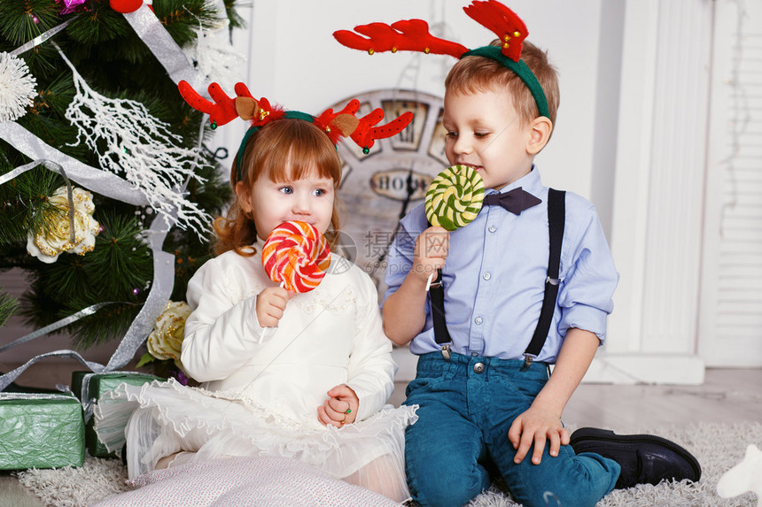 小女孩和男孩在驯鹿角吃棒糖两个有趣的小孩手里拿着美味的糖果的画像圣诞节和新年的概念快乐的孩子和家人期待图片