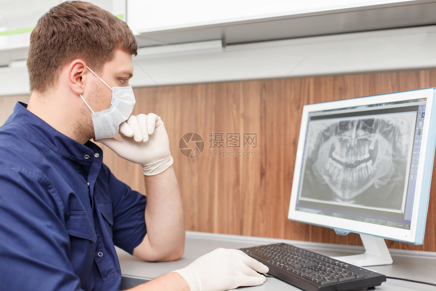 格开朗的男牙医正坐在桌旁使用他正在分析人类牙齿的X光片男人正认真地看着屏幕他戴图片