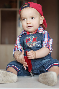 穿着牛仔裤工装裤和红帽的快乐幼儿男孩拿着平鼻钳室内肖图片