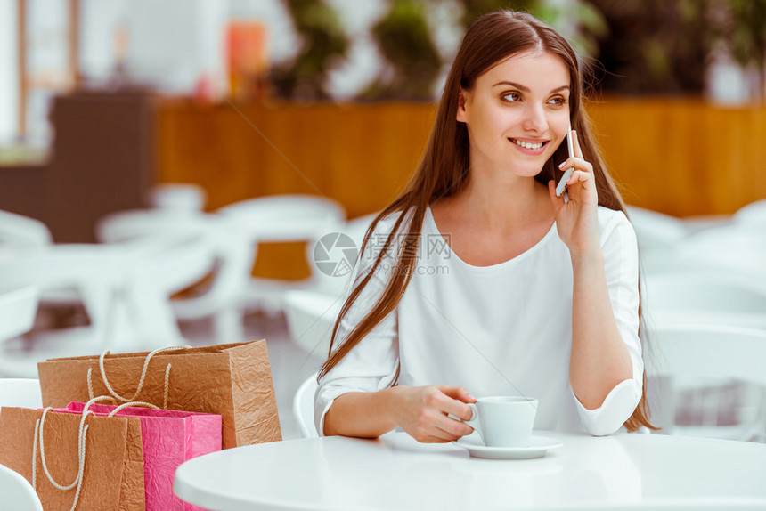 穿着白色上衣的年轻美女在买东西后在咖啡厅休息时用手机聊天喝咖啡和微笑图片