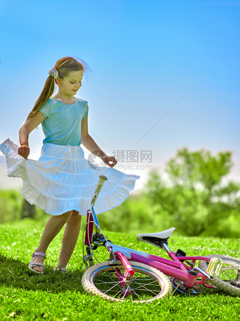 骑自行车的女孩穿着白裙子的女孩在自行车附近很高兴女孩旋转蓝色的天空和背景上的绿树图片