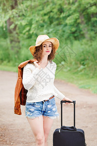 身穿白色毛衣蓝色牛仔短裤草帽背着旅行袋站在森林野乡公路上的美丽高图片