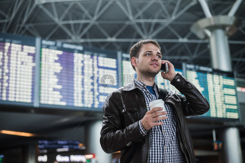 机场的年轻旅行者在时间表板前用电话交谈时微笑着图片