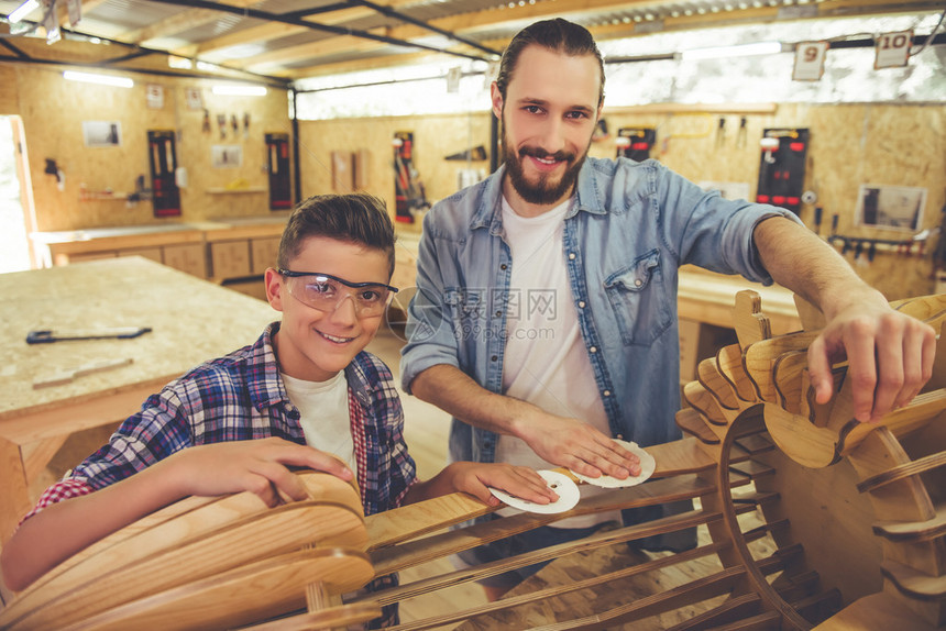 身戴保护眼镜的英俊十几岁的木匠和他父亲在车间与木材和砂纸一起工作时看着照相图片