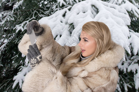 穿着毛皮大衣的年轻女子站在雪盖着公园她用电图片