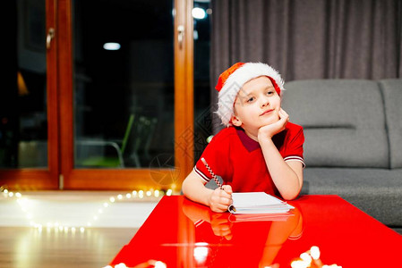 沉思的小男孩给圣诞老人写了一封信戴着红色圣诞老人帽背景图片
