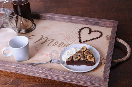 一块巧克力蛋糕配香蕉法式咖啡机和木托盘上的奶油罐咖啡豆的心文字图片