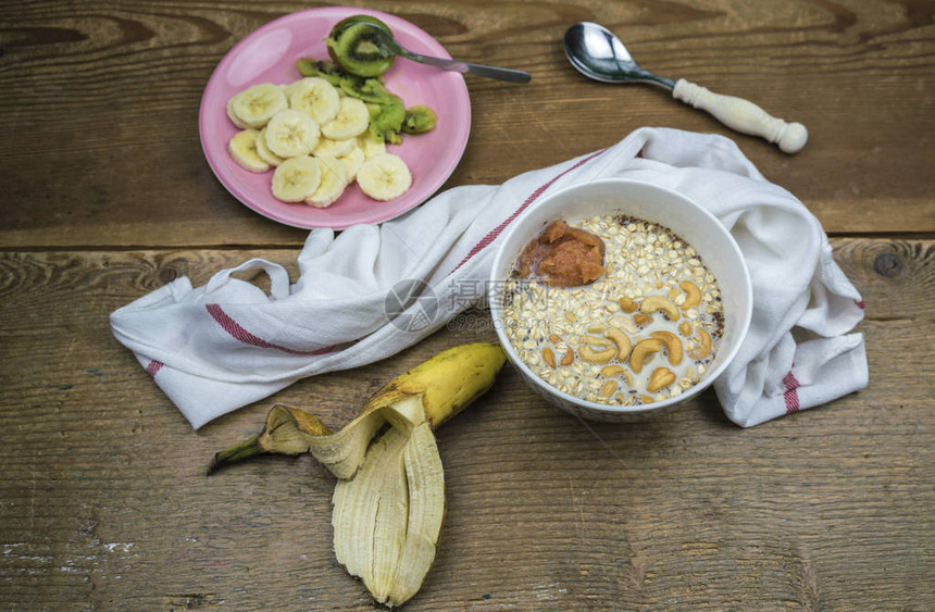 木桌上白碗牛奶和腰果燕麦片香蕉和猕猴桃图片