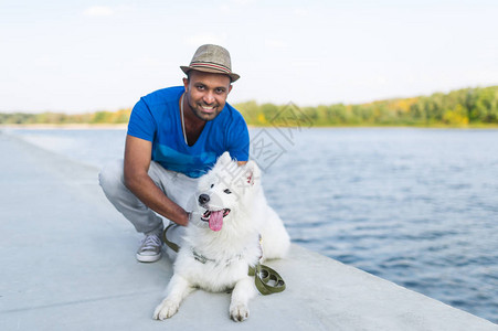 快乐的年轻斯里兰卡男子与她的狗白色萨摩耶坐在河边图片