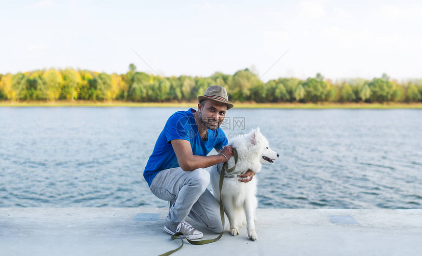 快乐的年轻斯里兰卡男子与她的狗白色萨摩耶坐在河边可爱的年轻斯里兰卡男人抱着图片