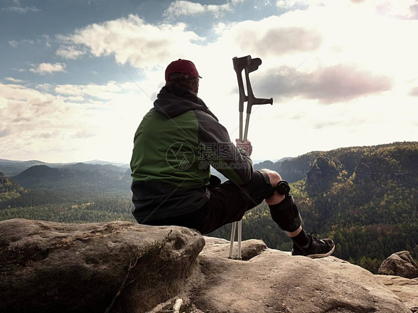 疲惫的游客用药拐杖和断腿固定在山顶上的防盗器中Valleybellow坐在绿色风衣和红色棒球帽的男人锋图片