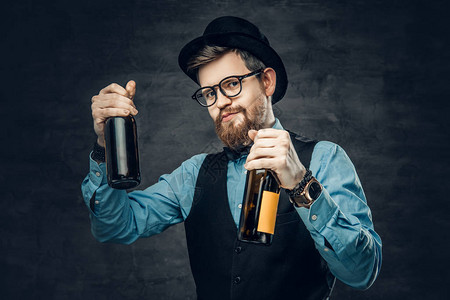 穿着蓝色衬衫优雅的腰衣和顶帽的长胡子男肖像中了两个手工艺啤酒瓶图片