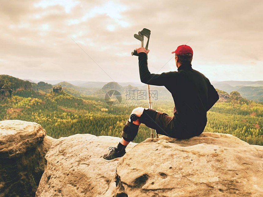 疲惫的游客用药拐杖和断腿固定在山顶上的防盗器中Valleybellow坐着穿黑色运动衫和红色棒球帽的男人锋图片