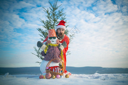 男子在多云的天空上与雪雕合影圣诞老人帽子和服装的惊奇时髦戴眼镜的雪人和头上的水桶圣诞树和新年礼物在雪地背景图片