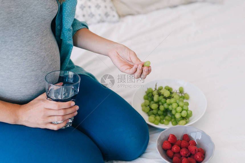 年轻孕妇用水杯在床上吃水果的照片以作图片