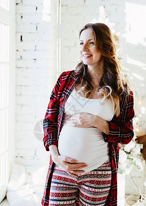 美丽的孕妇快乐地站在窗边在明亮的白色房间触摸她的肚子图片