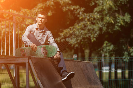 穿着时髦的衬衫运动鞋和裤子的笑年轻男子的肖像与滑板坐在滑冰公园中阳光图片
