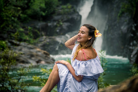 穿着裙子的漂亮女孩和坐在瀑布边坐着的头发中的Hibiscus花朵图片