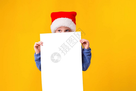 可爱的小女孩在圣诞老人帽子与黄色背景上的白色空白纸页图片