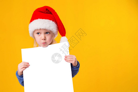 可爱的小女孩在圣诞老人帽子与黄色背景上的白色空白纸页图片